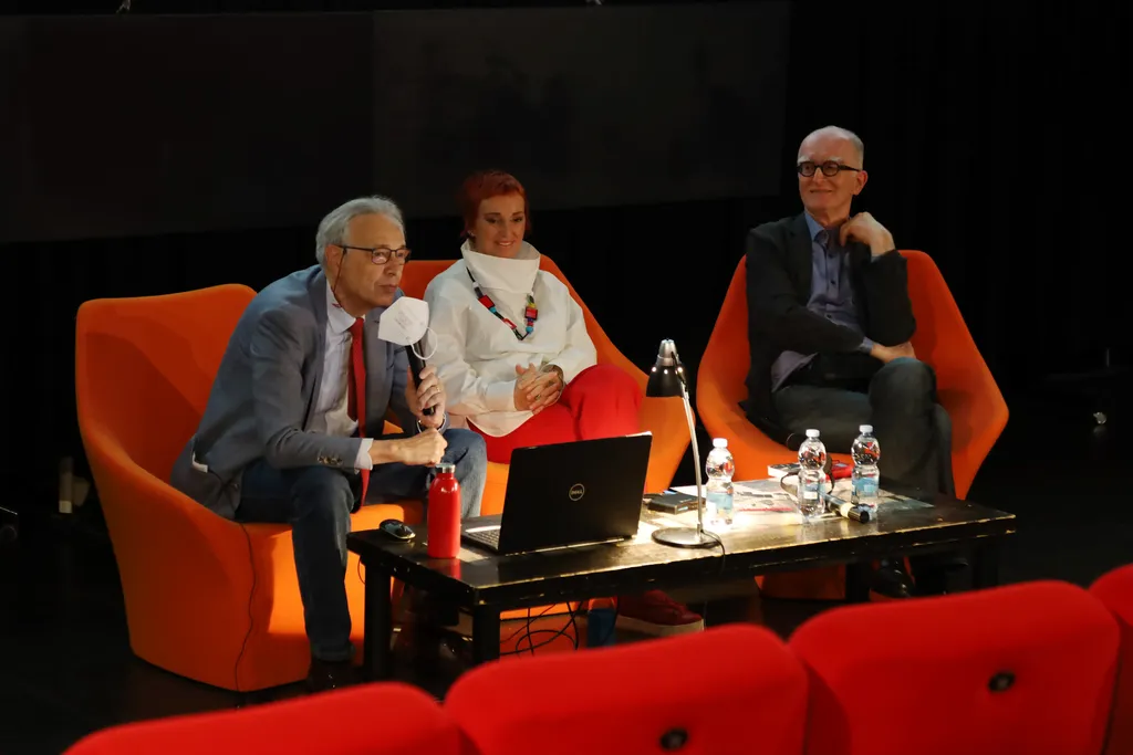 Photo of the round table event with three interlocutors. A laptop computer, a table lamp and four bottles of water are placed on the table.