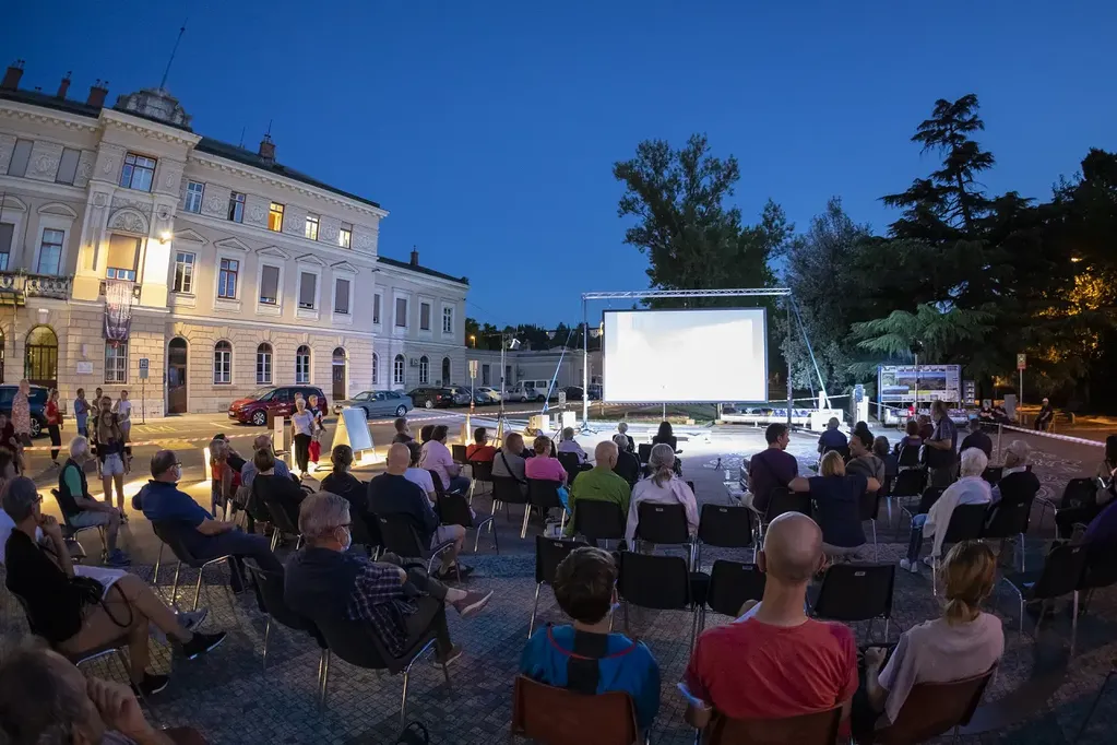 The photo shows the event at Trg Evrope, in front of the Nova Gorica railway station. A group of visitors are sitting on chairs and are closely watching what is happening on the screen.