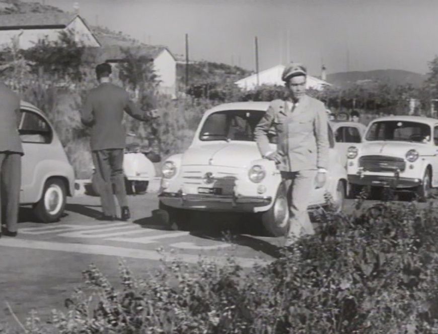  sign. Two gentlemen look at the first car while a third gentleman, dressed in uniform, walks past them.