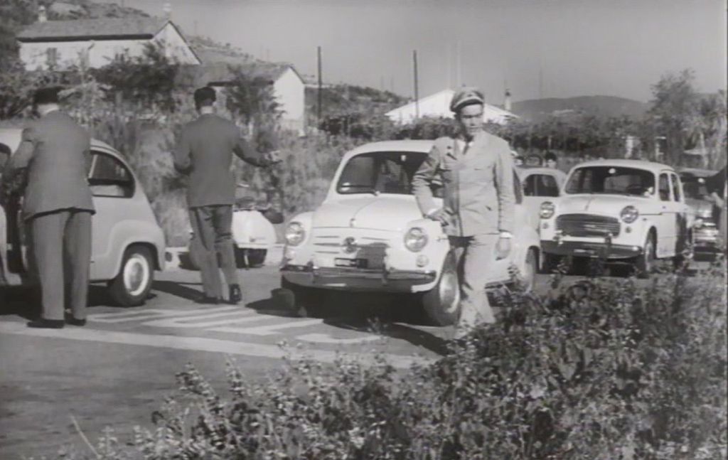 Črno-bela starinska fotografija staromodnih avtomobilov, ki so postavljeni pred znakom \&quot;STOP\&quot;. Dva gospoda si ogledujeta prvi avtomobil, medtem ko tretji gospod, oblečen v uniformo, gre mimo njih.