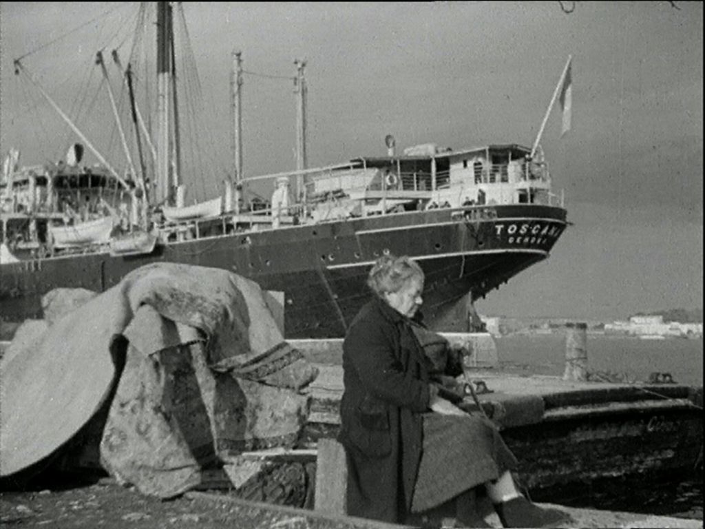 Vintage black and white photo shows an elderly lady sitting on a pier. In the background is an Italian passenger ship.