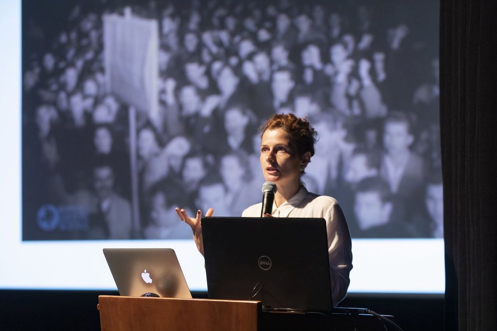 Photo of a female lecturer lecturing to an audience. She is wearing a white shirt and has short curly hair. He is holding a microphone in his hand.