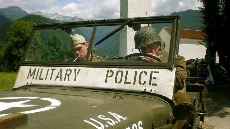 Fotografija iz filma z naslovom \&quot;Nočemo belega kruha\&quot; prikazuje dva ameriška vojaka, ki si prižigata cigareti in sta oblečena v uniformah iz druge svetovne vojne. Oba vojaka sedita v džipu, ki ima napis \&quot;Military police\&quot;.