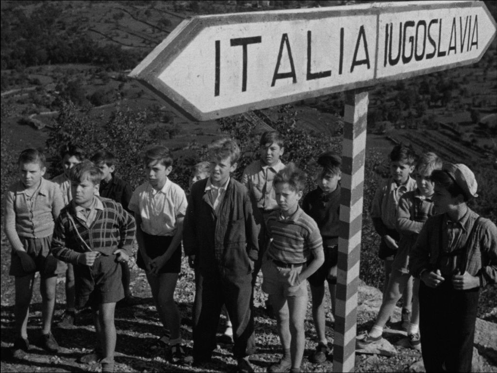 Una foto in bianco e nero dal film \&quot;Cuori Senza Frontiere\&quot; che mostra un gruppo di bambini in piedi davanti al cartello Italia, Jugoslavia.