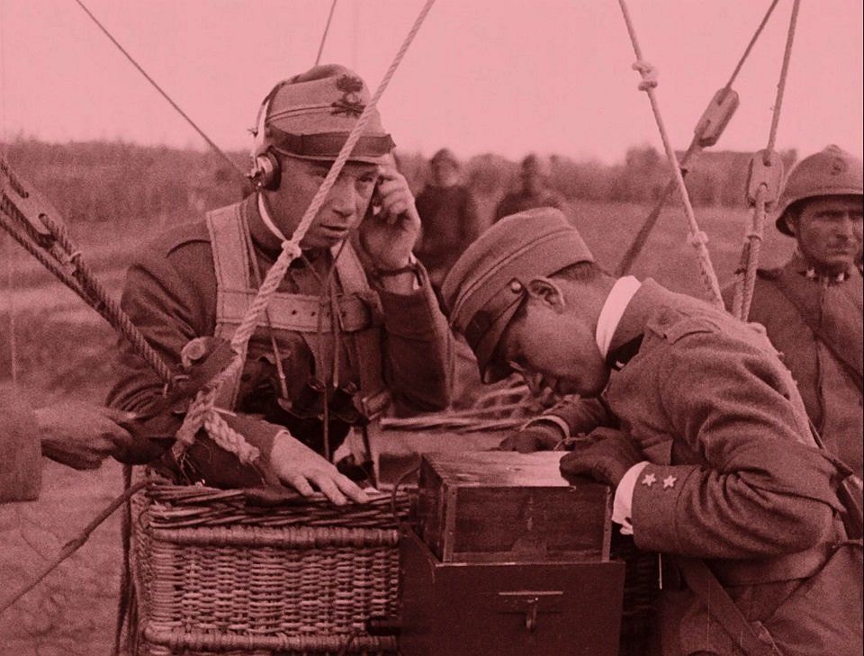 Vintage photo of two Italian soldiers. The soldier on the left is standing in a basket and listening attentively to a message on the radio. The soldier next to him looks intently at the wooden box.