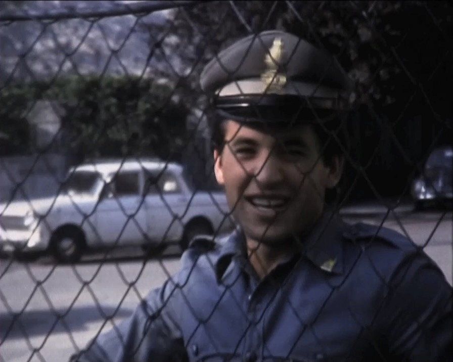 Foto dal film \&quot;La confessione del contrabbandiere\&quot; di un giovane sorridente in uniforme blu. Di fronte è posta una recinzione.